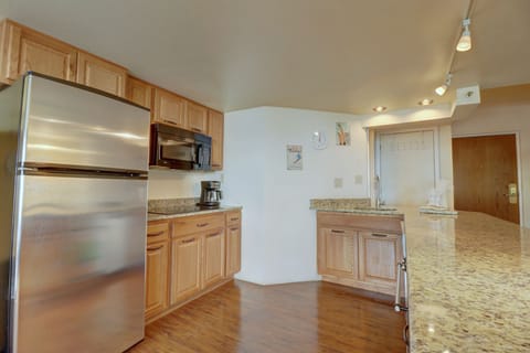 Modern kitchen with wooden cabinets, stainless steel appliances including a refrigerator and microwave, granite countertops, hardwood floors, and track lighting. Door and clock visible in background.