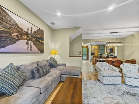 A living room with a gray sectional sofa, patterned chairs, wooden coffee table, wall art of a lake scene, and a view into a kitchen and dining area with a light fixture and brown dining chairs.