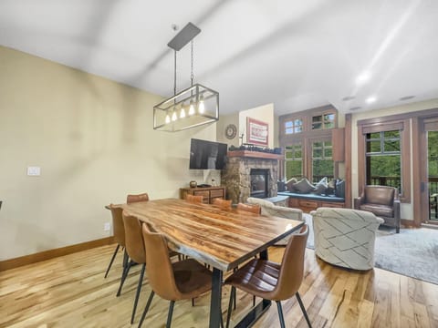 A modern dining room with a wooden table, six brown chairs, a large rectangular light fixture, and an adjacent living area with a fireplace, cozy seating, and large windows.