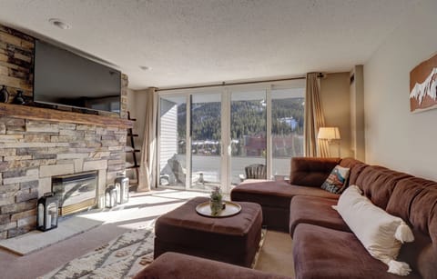 A cozy living room with a stone fireplace, a large wall-mounted TV, a brown sectional sofa, and large sliding glass doors opening to a balcony with a scenic mountain view.