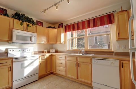 A bright kitchen with wooden cabinets, a white stove, microwave, dishwasher, and a large window featuring striped red curtains. Countertop includes a sink and various small appliances. Floor is tiled.
