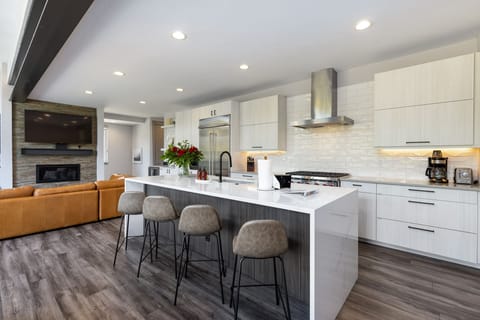 Modern kitchen with white cabinets, a large center island with four bar stools, stainless steel appliances, and wood flooring. Living area with a brown couch and fireplace is visible in the background.