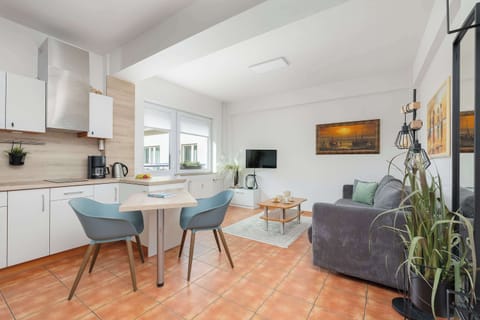 A modern kitchen with white cabinets, a gray couch in the living area, and a dining table with blue chairs. A plant is visible in the foreground.