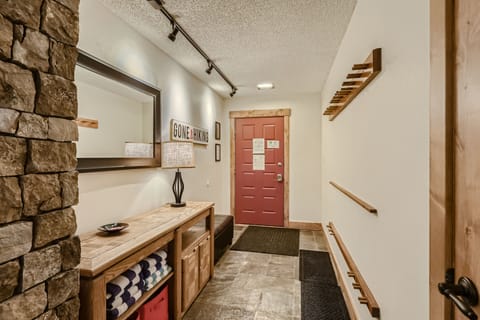 A hallway with a red door, tiled floor, stone accent wall, wooden bench, wall mirror, lamp, and a "Gone Fishing" sign. Shelves holding towels and boxes are below the bench. Track lighting on ceiling.