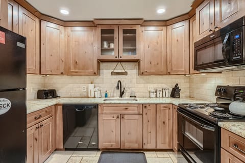 A kitchen with wooden cabinets, granite countertops, a black dishwasher, refrigerator, microwave, and stove. The sink is centered under cabinets with glass doors. Illuminated by ceiling lights.