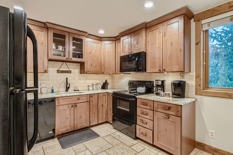 A kitchen with wooden cabinets, a black refrigerator, black stove and microwave, dishwasher, coffee maker, and a window with a view outside. There’s a mat in front of the sink and tiled flooring.