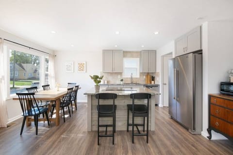 Open-concept kitchen & dining: sleek island with two bar stools faces a wood table for six by bright windows. Gray cabinets, stainless fridge, and wood floors create a harmonious flow throughout the space. 🍽️🏡✨