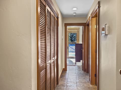 A narrow hallway with tile flooring, wooden louvered doors on the left, doorways on the right and at the end, leading to a room with a view of an outdoor deck.