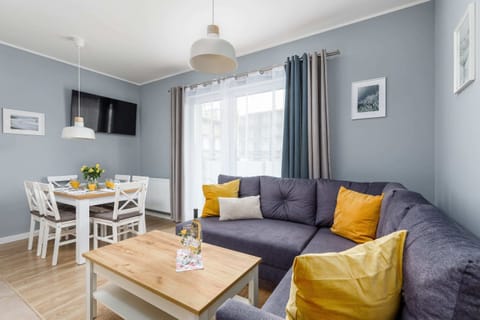 A cozy living room with a gray sofa adorned with colorful cushions. A coffee table and a small dining area in a bright, modern setting complete the space.