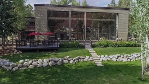 A modern building with large floor-to-ceiling windows, a wooden deck with red umbrellas, and a stone-bordered lawn with a pathway leading to the entrance. Trees surround the structure.