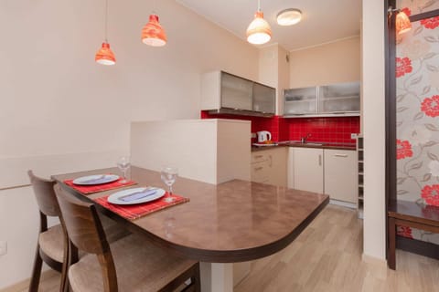 A dining area with a wooden table and chairs, adjacent to a modern kitchen featuring red tiles and pendant lights.