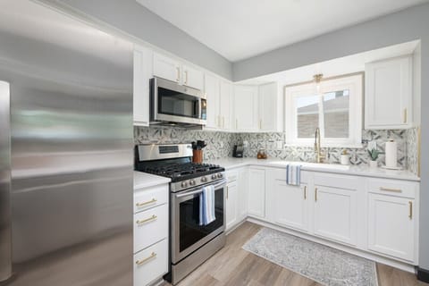 Kitchen features stainless steel appliances and gas stove   - White cabinets with modern gold pulls and tile design   - Large window provides natural light for cooking and meals