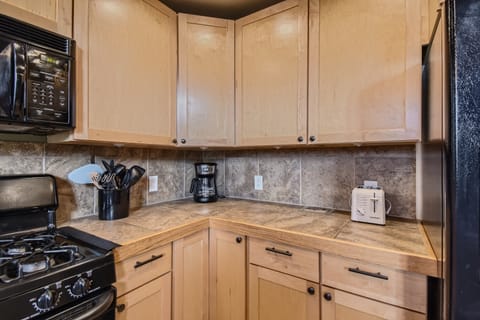 A kitchen with wooden cabinets, a stovetop with black appliances, a coffee maker, a toaster, and a utensil holder on a tiled countertop with a tiled backsplash.