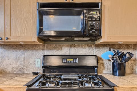 A kitchen with a black microwave mounted above a black gas stove and oven. Utensils in a holder are on the right side on a tiled backsplash.