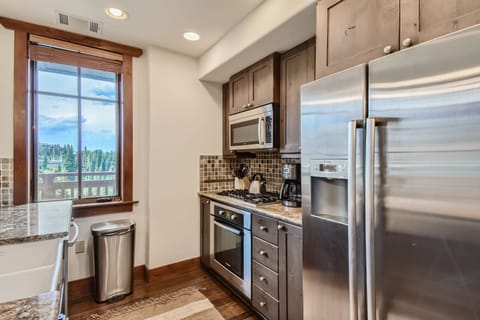 A modern kitchen with stainless steel appliances, including a refrigerator, oven, and microwave, wood cabinets, a tiled backsplash, and a window with a view of nature.