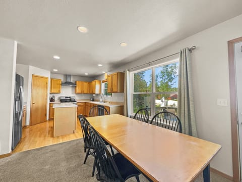 A wooden dining table with black chairs is set in an open-plan kitchen and dining area. The kitchen has wooden cabinets, stainless steel appliances, and a window offering a view of trees outside.