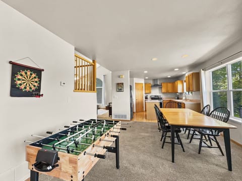 A living space with a foosball table, dartboard on the wall, dining table with black chairs, and a kitchen with wooden cabinets and stainless steel appliances in the background. Large window to the right.