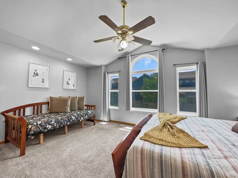 A bedroom with a ceiling fan, a daybed with patterned cushions, and a large bed with a striped cover. Windows with white trim let in natural light. Two framed pictures hang on the wall above the daybed.