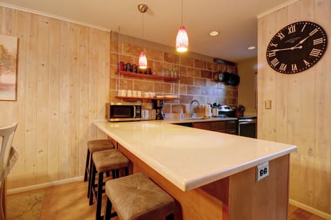 A cozy kitchen with wooden walls, a large wall clock, and a white countertop with three bar stools. Pendant lights hang above the counter, and kitchen appliances are visible in the background.