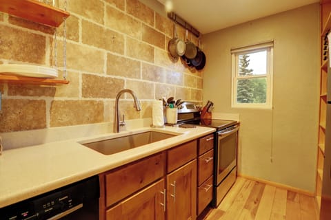 A kitchen interior with wooden cabinets, a stainless steel sink, an oven, a stove, and a window. Pans hang on the wall, and a stone backsplash adds texture.