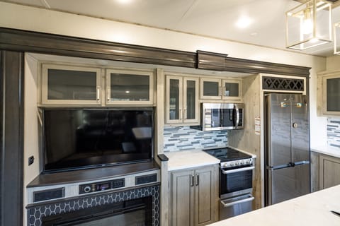 View of TV and Kitchen with three burner stove and residential refrigerator.