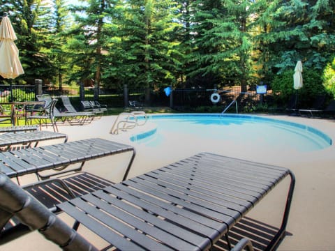 Outdoor swimming pool area with lounge chairs, umbrellas, and surrounding tall evergreen trees.