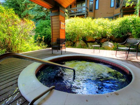 Outdoor hot tub surrounded by lush greenery and a wooden deck, with two black chairs positioned nearby.