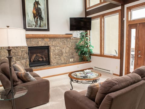 A cozy living room with a stone fireplace, two brown sofas, a round glass coffee table, a flat-screen TV, a large painting of a horse, a floor lamp, and a plant. Large windows offer outdoor views.