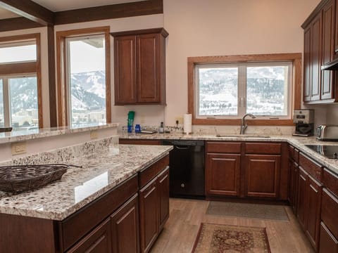 A kitchen with dark wood cabinets, granite countertops, a stainless steel sink, and large windows with a view of snowy mountains. The room includes various kitchen appliances and a woven basket on the counter.