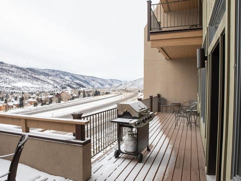 A snowy deck with a gas grill, tables, and chairs overlooks a mountainous landscape and a rail track in the distance.