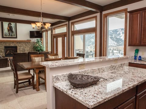 A modern kitchen with a granite countertop, a wicker basket, and dark wood cabinetry. Adjacent dining area with a wooden table and leather chairs, and a living room with a stone fireplace and mountain view.