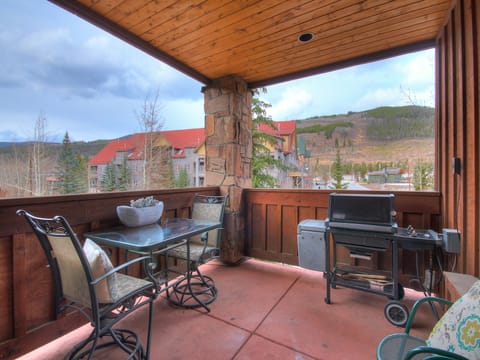 Covered balcony with a glass-top table, four padded chairs, a grill, and a view of a residential area and distant hills.