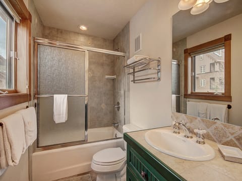 A bathroom with a bathtub and shower combo with sliding glass doors, a toilet, a sink with a green cabinet, a wall mirror, and a towel rack. The window provides natural light.