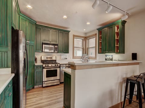 A kitchen with green cabinets, granite countertops, stainless steel appliances, a light wooden floor, and a small breakfast bar with a stool. It is illuminated by ceiling track lights.