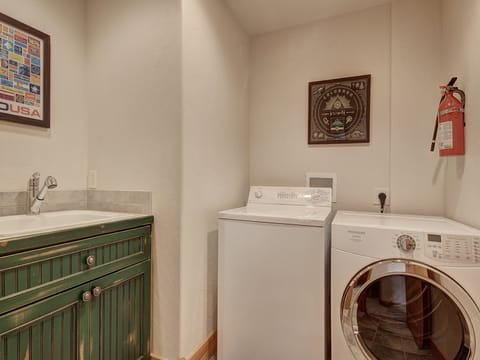 A laundry room with a green cabinet, a sink, a top-loading washing machine, a front-loading dryer, framed pictures on the wall, and a fire extinguisher mounted on the wall.