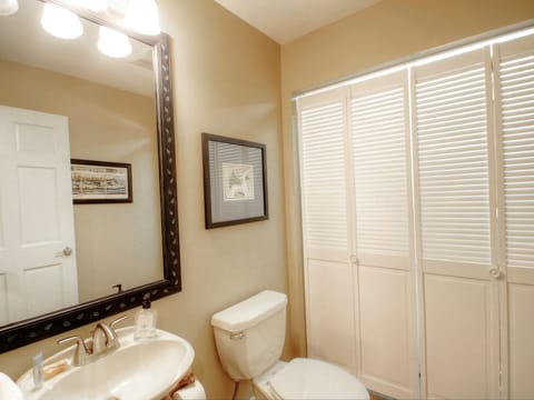 A bathroom with a toilet, sink, and mirror. Wall art is visible next to a window with closed blinds. A door is partly open on the left.