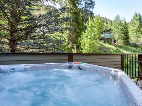 Hot tub on a wooden deck overlooking a lush, forested hillside with trees and a house in the background.