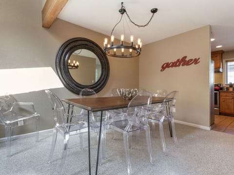 A dining area with a rectangular wooden table, clear plastic chairs, a round mirror, and a chandelier. The word "gather" is mounted on the wall. A kitchen is visible in the background.