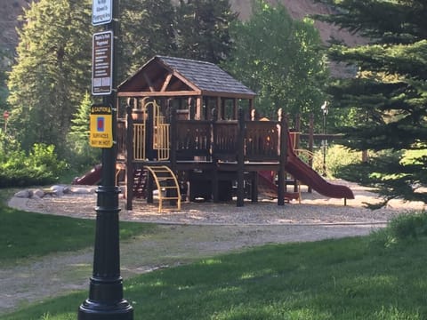 A wooden play structure with slides and climbing features is set in a forested park area. A sign near the structure warns that surfaces may be hot.