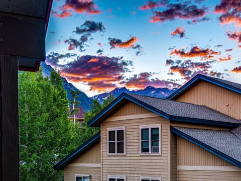 View of a multi-story building with beige siding and blue roof trim, set against a backdrop of green trees, snow-capped mountains, and a colorful sunset sky with scattered clouds.
