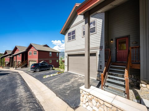 A row of modern, two-story houses with driveways and garages. The closest house is grey with a red door and stone base. A black car is parked in front of the second house. The sky is clear and blue.
