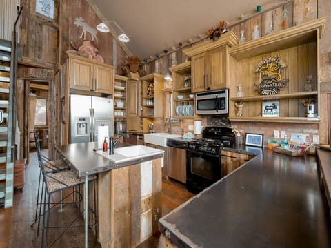 Rustic kitchen with wood-paneled walls, modern stainless steel appliances, a dark countertop island with stools, and open shelving. Decor includes small figurines and a vintage-style sign.