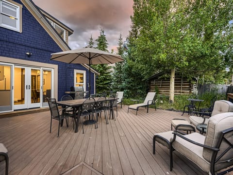 A wooden deck with outdoor furniture, including a dining table with chairs under an umbrella, cushioned lounge chairs, and a grill, is surrounded by greenery. The house exterior is blue with large windows.