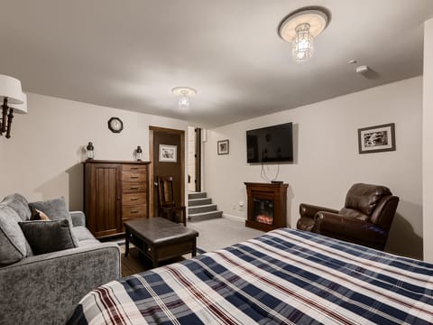 A cozy bedroom with a plaid bedspread, sofa, ottoman, wooden dresser, leather recliner, flat-screen TV, and a fireplace. Steps lead to a higher level at the back.