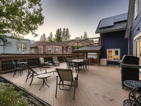 Outdoor patio with tables, chairs, an umbrella, and a grill, surrounded by a wooden fence. Houses and trees are in the background. The sky is clear, suggesting evening or late afternoon.