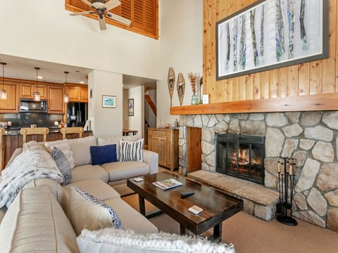 A cozy living room with a stone fireplace, beige sectional sofa, and wooden coffee table. Kitchen area with wooden cabinets is visible in the background. A framed artwork hangs above the fireplace.