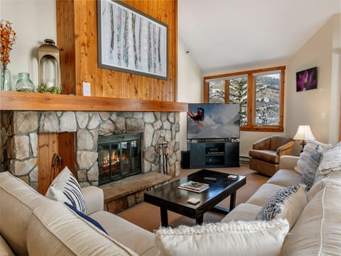 Cozy living room with a stone fireplace, wooden mantel, a flat-screen TV, comfortable seating, and large windows overlooking a snowy landscape.