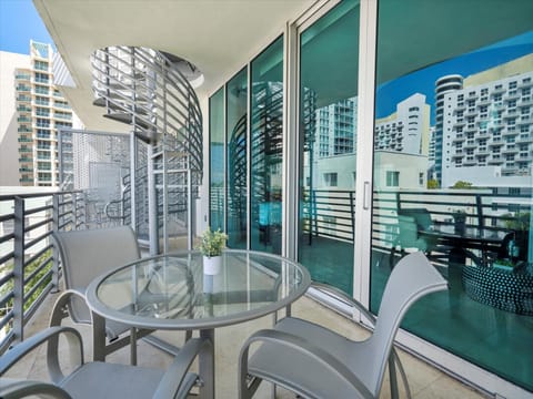 A modern apartment balcony with a glass table and four chairs. The balcony is enclosed by glass doors and features a spiral staircase. Tall buildings are visible in the background.