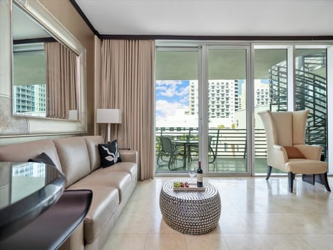 Luxurious living room with a beige sofa, mirror, round coffee table, and armchair near patio doors leading to a balcony. The view includes several buildings and a partly cloudy sky.