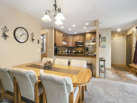 A dining area with a wooden table and six chairs, adjacent to a kitchen with wooden cabinets and stainless steel appliances. The room has beige walls and a tile floor near the entrance.